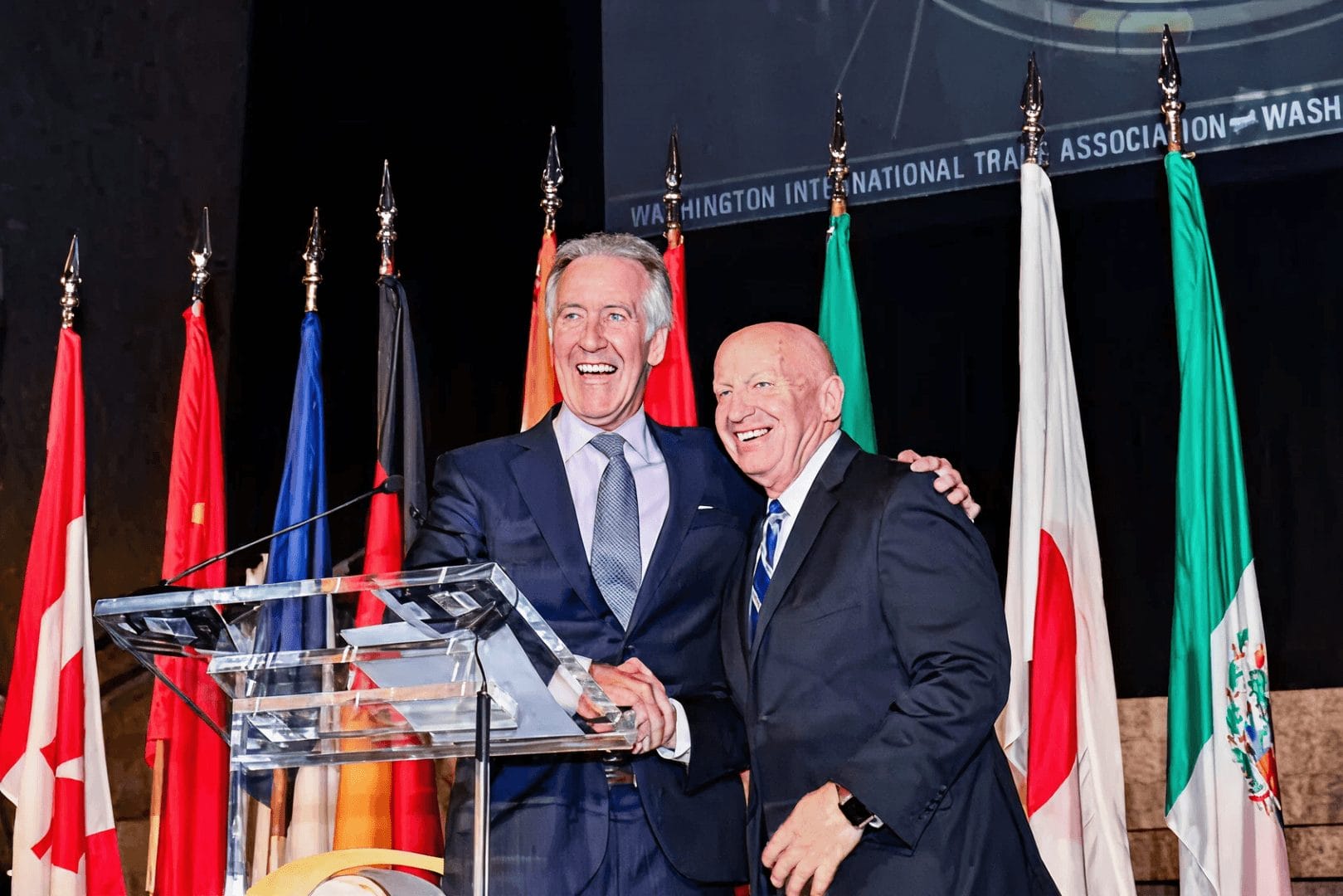 Two speakers shaking hands at a podium during a WITA event, with international flags and a Washington International Trade Association banner in the background