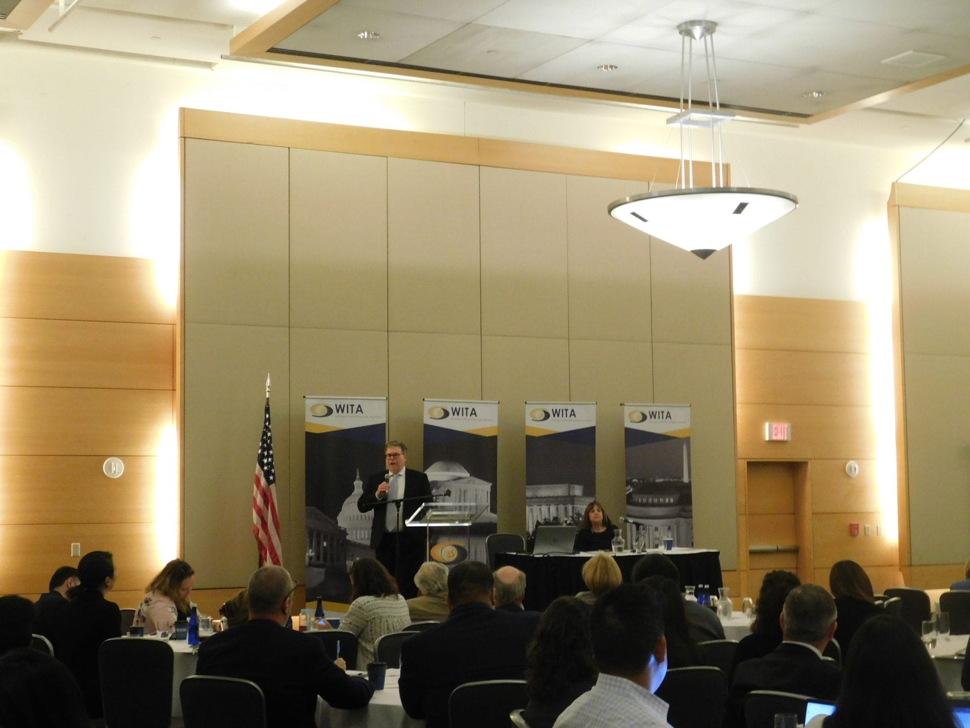 A speaker addressing the audience from a podium in front of WITA banners during a conference session